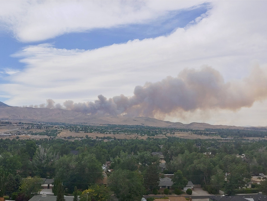 An aerial view of smoke billowing over buildings and vegetation with mountains in the background.